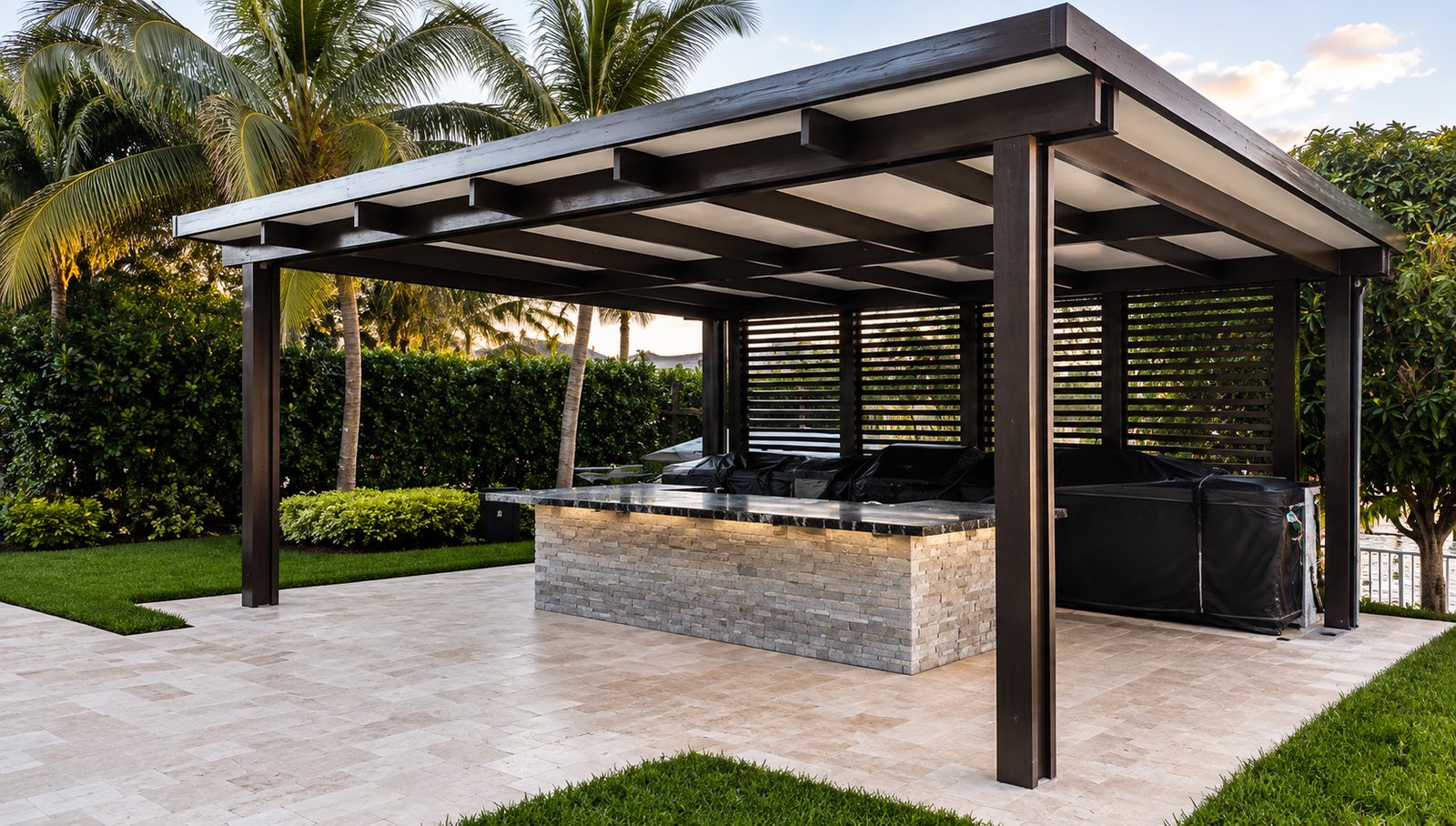 Dark bronze free-standing aluminum patio cover over a stacked-stone outdoor kitchen on travertine paving